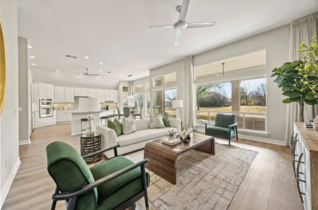 Large living room and kitchen staged with a large light grey sofa with green and sliver pillows, two velvet green chairs with dark wood frames, kitchen island with no back barstools.