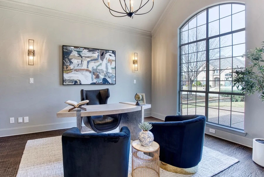 Home office staged with a modern desk, a black leather desk chair, two blue velvet barrel chairs, white rug and abstract art