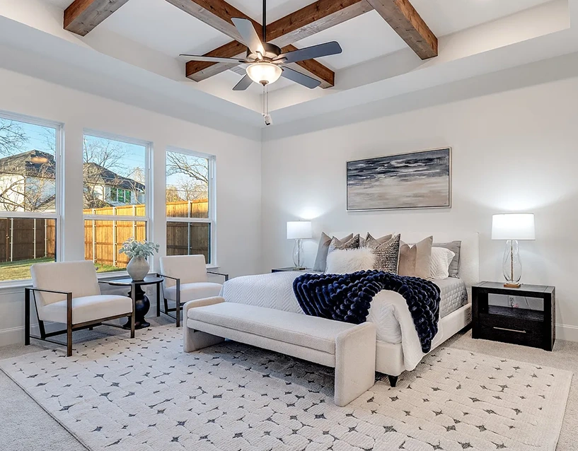 A beautiful staged bedroom in plano texas with a large white upholstered bed, grey and white pillows, a white upholstered bench, two white chairs with metal frams and two dark wood nighstands
