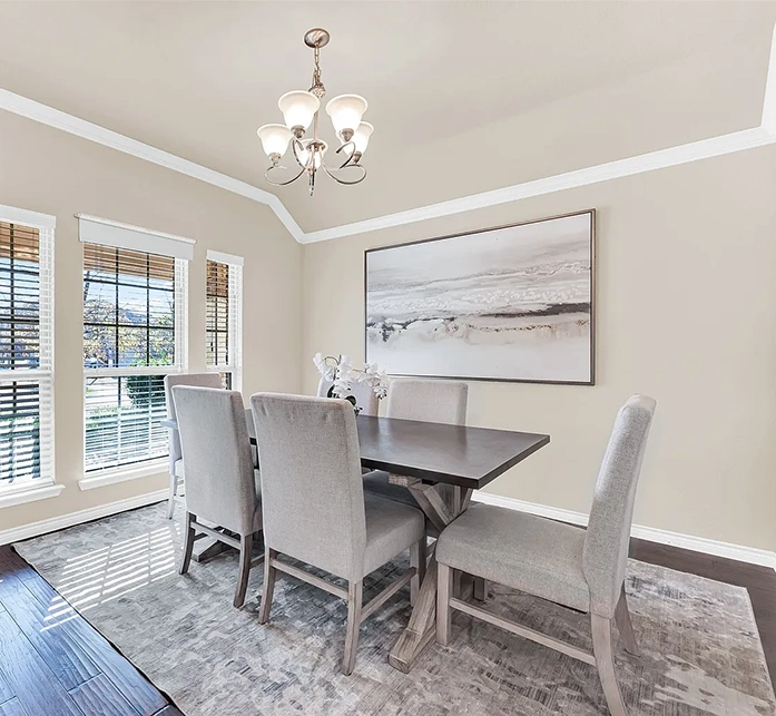 Dining room staged with a rectangular table, six high back grey velvet chairs, grey area rug and large artwork