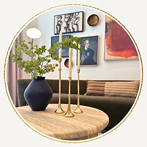 Close up of a coffee table staged with a black vase and faux greenery, three candle sticks and a gallery wall of artwork behind the sofa in the background