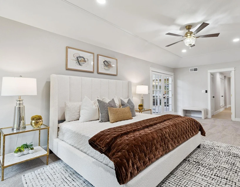 Staged primary bedroom showcasing a high-back cream tufted bed, a rich brown velvet throw blanket, glass nightstands, and a crystal-accented ceiling fan
