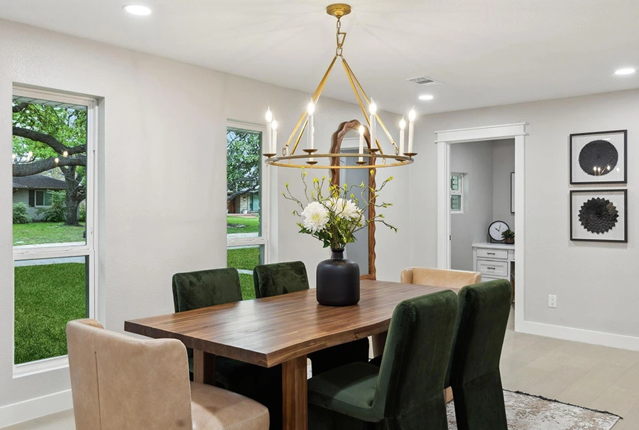 Staged dining room with a warm wood table, deep green velvet and tan leather chairs, a gold candle-style chandelier, and large bright windows