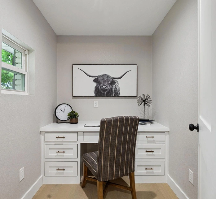 Staged pocket office nook with a built-in white desk, a striped upholstered desk chair, and a black-and-white highland cow framed art print