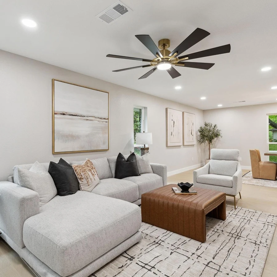 Staged living area or game room with a light grey sectional sofa, black accent pillows, a cognac leather ottoman coffee table, and a large multi-blade ceiling fan