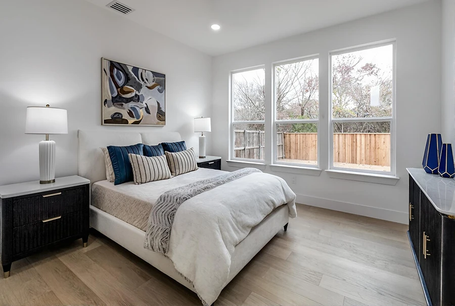 Staged secondary bedroom featuring a white upholstered bed, blue and striped accent pillows, black nightstands with gold pulls, and large windows looking out to a wood fence