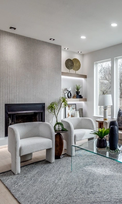 Vertical view of a staged living room seating area with two white bouclé chairs, a glass coffee table with black vases, and a fluted grey fireplace