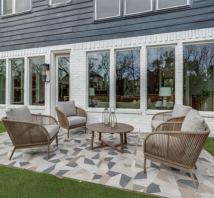 Staged outdoor patio featuring a patterned geometric tile floor, four woven wicker armchairs with grey cushions, and a round wood coffee table against a white brick exterior