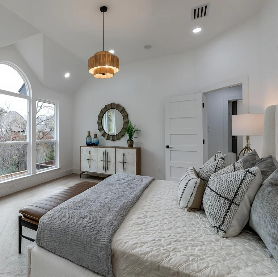 Staged primary bedroom shot from the side showing a white tufted bed with plush grey pillows, a grey quilted throw blanket, a brown leather bench, and a tiered wood chandelier