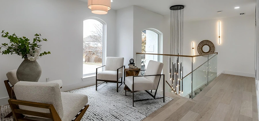 Staged second-floor landing with two white accent chairs on a textured black-and-white rug, a glass railing overlooking a staircase, and a dramatic, multi-light black pendant chandelier