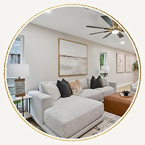 Circular vignette of a staged modern living room showcasing a large grey sectional sofa with black throw pillows, a tan leather tufted ottoman coffee table, and minimalist abstract wall art