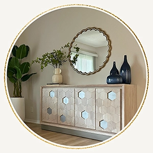 Circular vignette of a staged entryway highlight featuring a light wood sideboard console with a geometric hexagon pattern, dark blue ceramic vases, a round scalloped gold mirror, and a tall potted plant