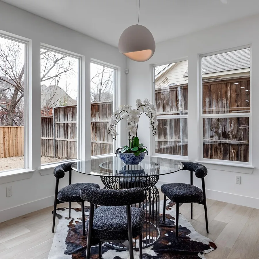 Staged breakfast nook featuring a round glass-top table, dark textured chairs, an orchid centerpiece, and a cowhide rug in front of large windows