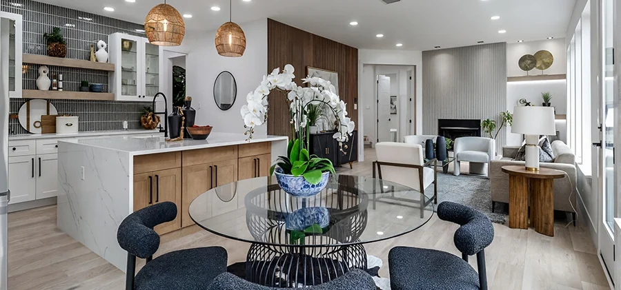 Staged open-concept main floor looking from a glass breakfast table past a kitchen island into a modern living room with a white-ribbed fireplace