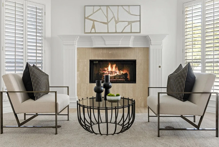 Seating area by a fireplace with two modern white chairs with black pillows, a black and glass round coffee table and white with gold artwork