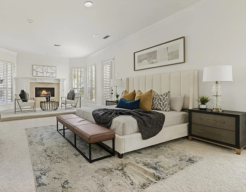 Staged primary bedroom with a large white upholstered bed, plush bedding, leather bench and two modern white chairs with grey pillows