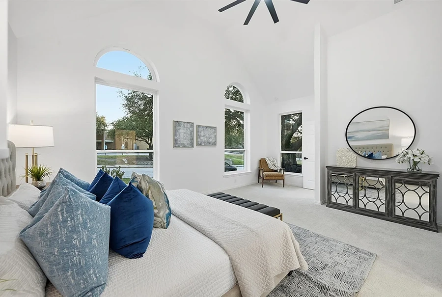 Large primary bedroom room with large arched windows, a white bed with blue pillows, a mirror front dresser and round mirror