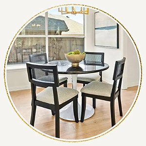 A circular photo of a bright dining area featuring a round glass table, four black wood chairs and a small gold light hanging above.
