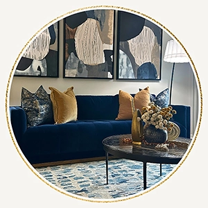 A circular photo showing a navy blue velvet sofa, tan and blue pillows, a round glass coffee table and three large abstract pieces of art hanging above the sofa.