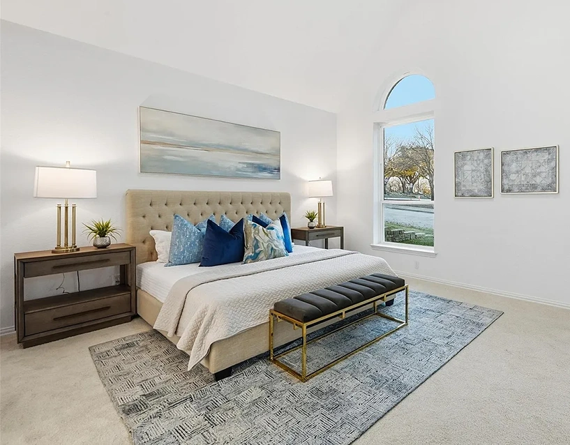 Luxury staged bedroom with a large tan upholstered bed, white bedding with lots of blue pillows, a grey leather bench and two wood nighstanbds with gold lamps