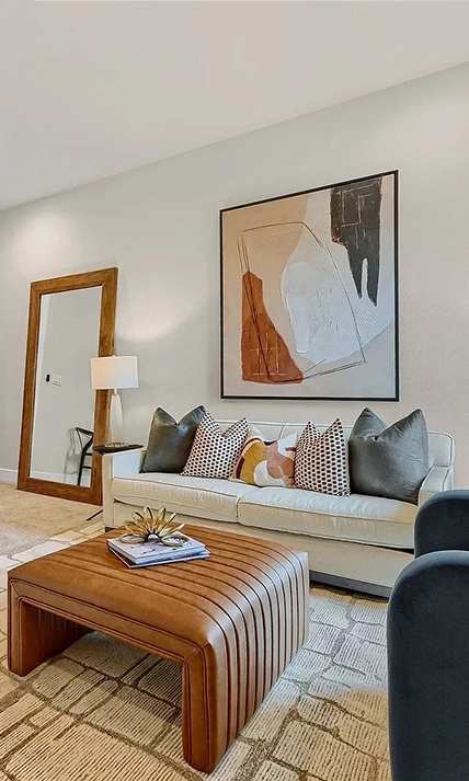 A modern seating area with a tan channel-tufted sofa, cognac leather ottoman and a large piece of abstract art hangs above the sofa.