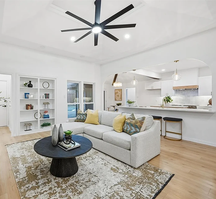 Wide angle view of living area opening up to a bright kitchen, living room featuring a light grey sofa with chaise lounge, black round coffee table and bookshelves with modern accessories.