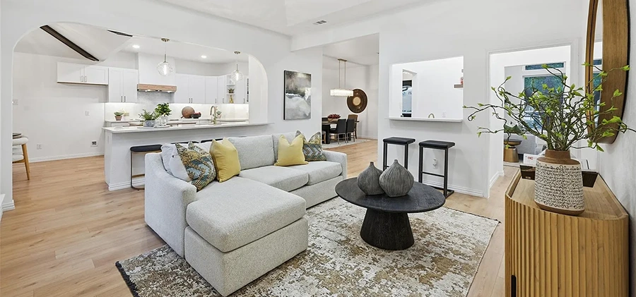 A bright living room featuring a light grey sofa with chaise lounge, black round coffee table, white kitchen in the background with low back black barstools
