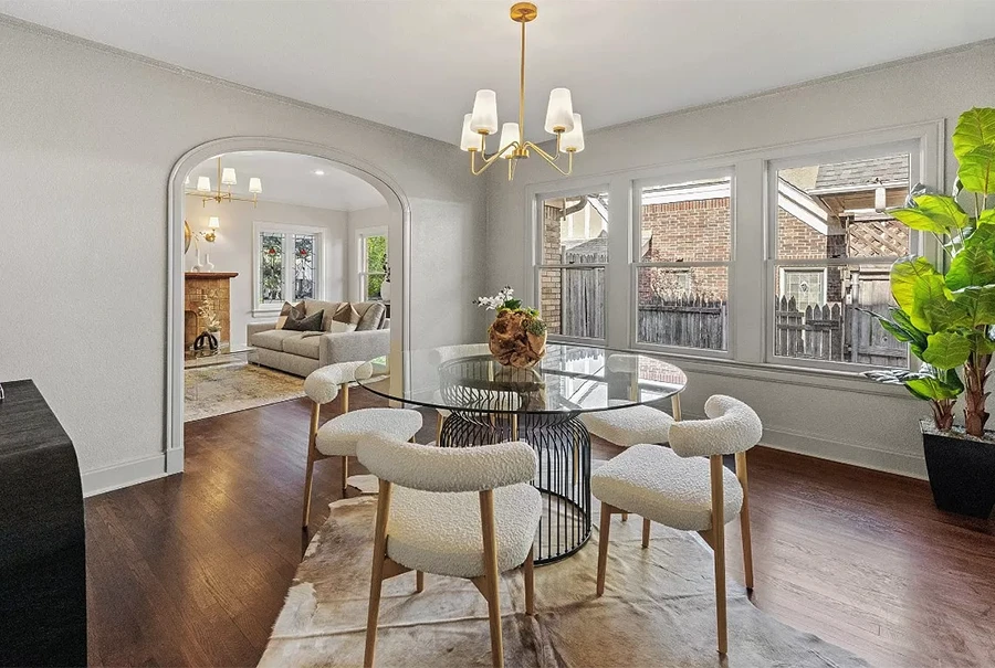 A large photo of a dining room with a glass-top wire-base table, white bouclé chairs, and a view through a classic archway into a living room with a fireplace.
