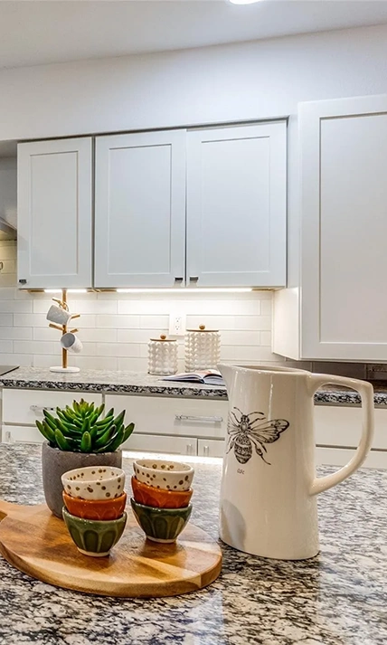 A close-up of a granite countertop featuring a white pitcher with a bee illustration, small colorful bowls, and white shaker-style cabinets.