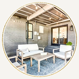 A circular photo showing a grey brick patio with a white modern outdoor sofa, two matching armchairs, and a wooden coffee table under string lights.