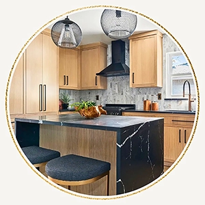 A circular photo of a kitchen featuring light wood cabinetry, a black marble island with a waterfall edge, and black barstools.