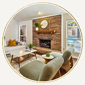 A circular photo of a cozy living room with a red brick fireplace, a rustic wood mantel, and two green mid-century modern armchairs.