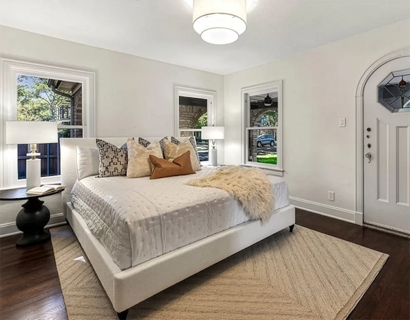 A bright bedroom with a white upholstered bed, neutral linens, and a tan throw rug, highlighting the natural light from two large windows.