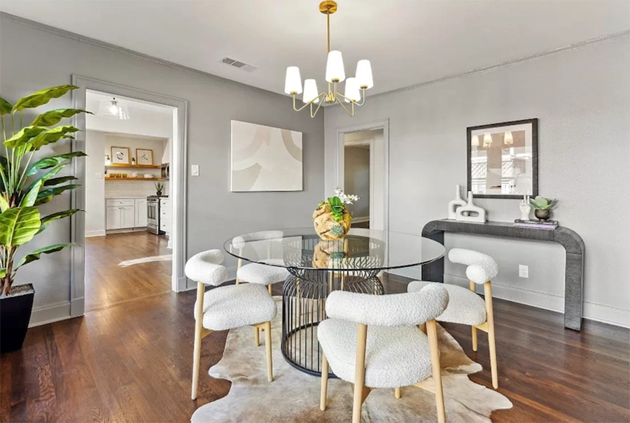 A round glass dining table surrounded by four white bouclé chairs with light wood legs, positioned under a modern brass chandelier in a room with grey walls.
