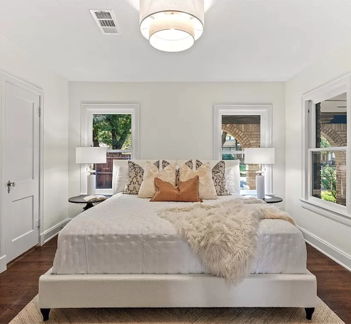 A plush white bed with tan and patterned pillows, flanked by classic wooden windows and modern bedside lamps.