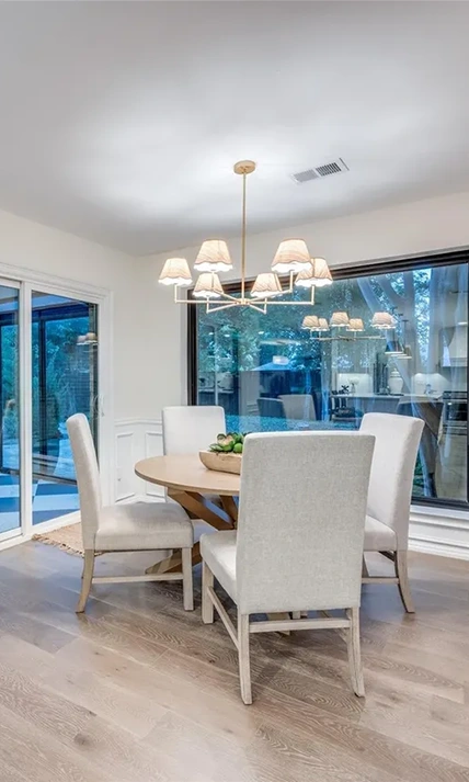 Staged dining space with a round wooden table, four cream high-back chairs, and a gold multi-shade chandelier near a sliding glass door
