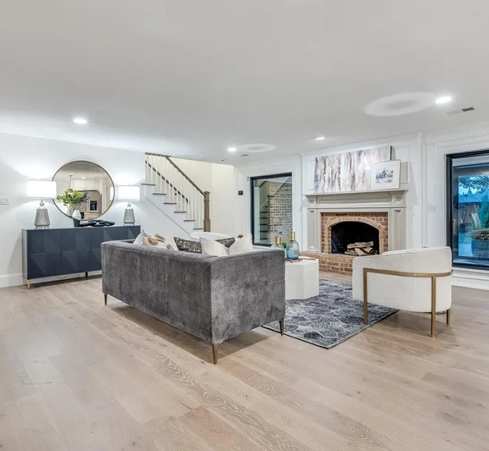 View from behind a grey velvet sofa showing a staged living room with a brick fireplace, white armchair, and dark blue buffet cabinet