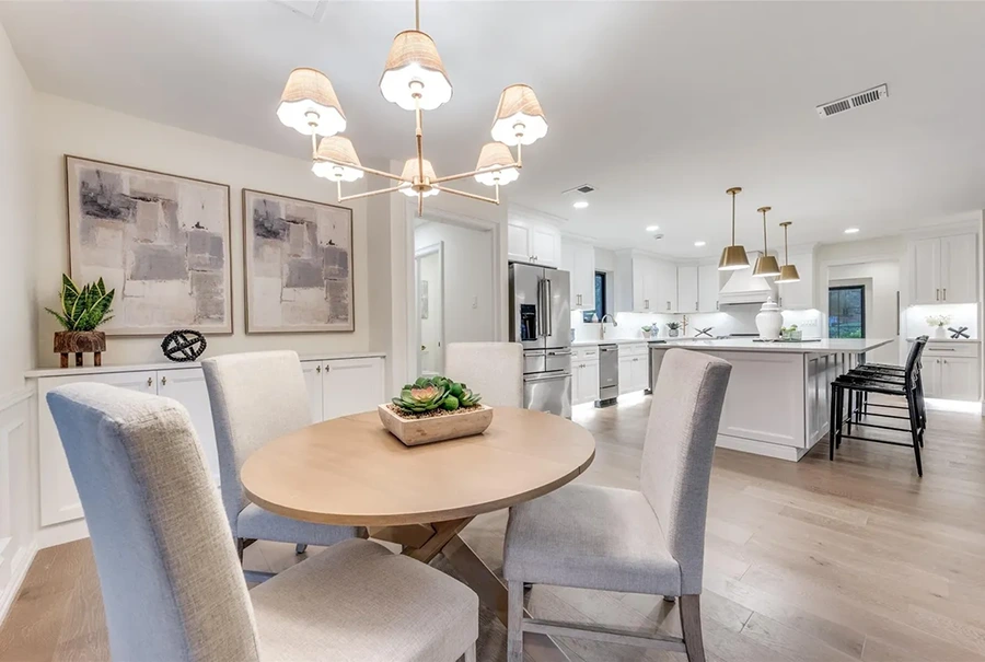 Staged casual dining area with a round wooden table, cream upholstered chairs, and a white kitchen visible in the background