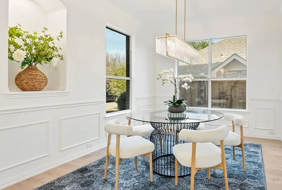 Staged bright dining room featuring a round glass-top table with a black wire base, four white textured sherpa chairs with wood legs, and a bold navy geometric area rug