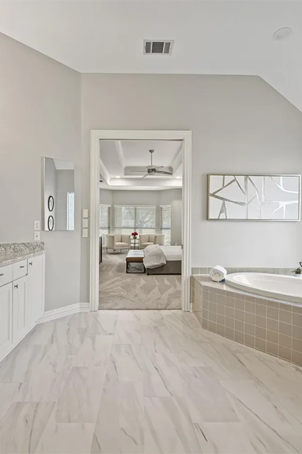 Spacious staged primary bathroom with marble-patterned floor tiles, a built-in soaking tub with beige tile accents, and an open doorway leading into the primary bedroom