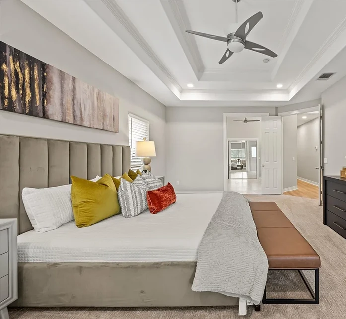 Staged primary bedroom showcasing a high-profile grey velvet vertical tufted headboard, accented with chartreuse and rust velvet pillows, beneath a large tray ceiling with a modern fan