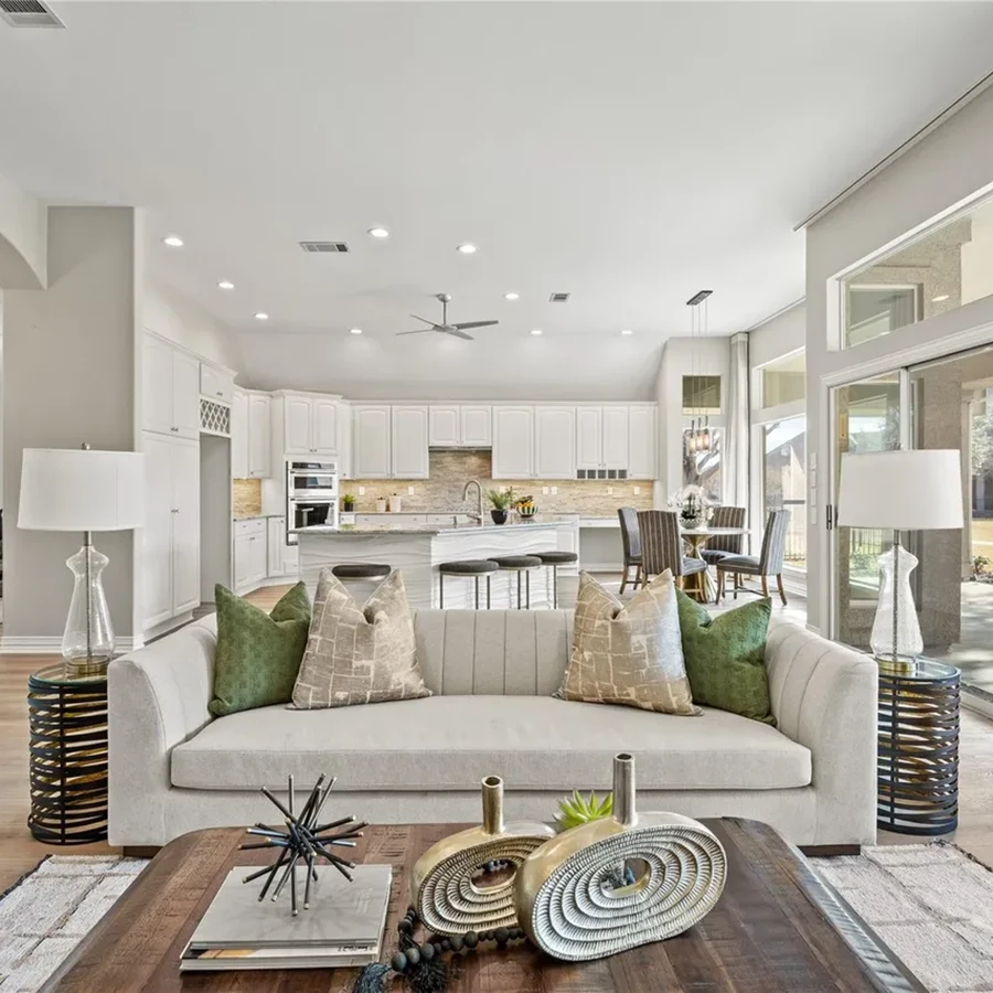 Open-concept staged living room looking past a textured cream sofa with forest green accent pillows toward a large white kitchen island and distant dining table