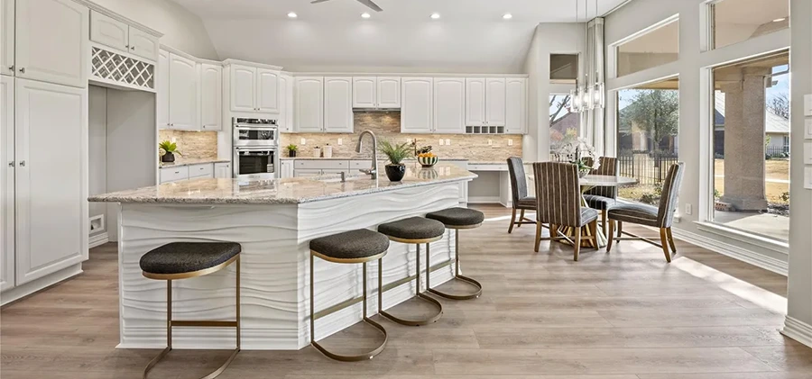 Staged modern kitchen highlighted by a long island with a white wavy textured facade, granite countertop, and four dark upholstered barstools with gold frames