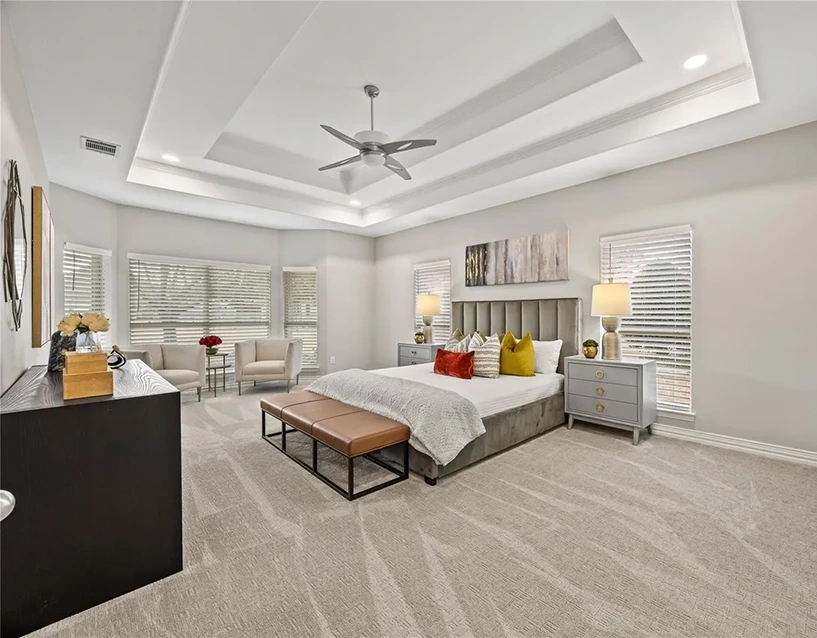 Staged primary bedroom featuring a grey velvet tufted headboard, a mix of amber and neutral accent pillows, a brown leather bench at the foot of the bed, and grey three-drawer nightstands