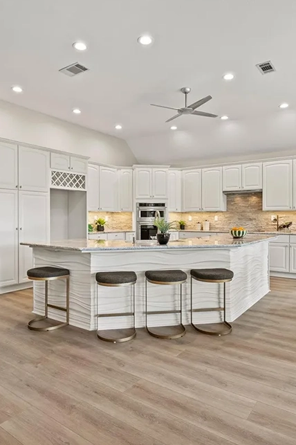 Staged spacious kitchen showing white cabinetry, a large curved island with a wavy textured white facade, granite countertops, and four dark upholstered barstools