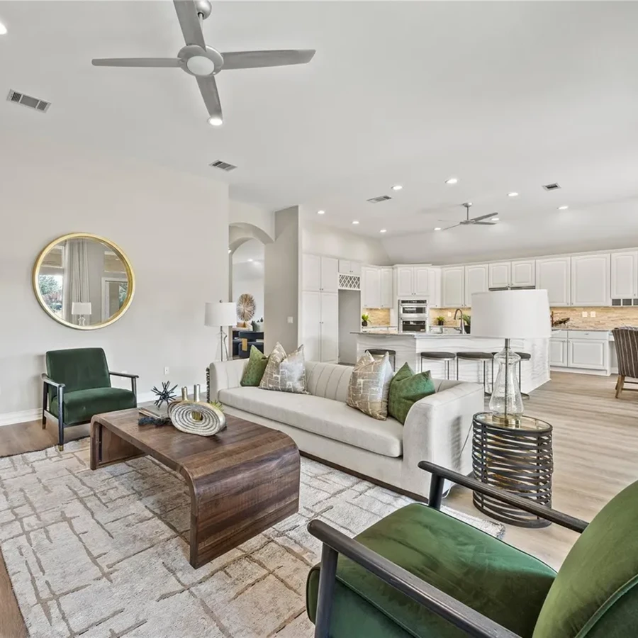 Angular view of a staged living space featuring a low-profile curved wood coffee table, a beige sofa with green patterned pillows, forest green armchairs, and a peek into the white kitchen behind