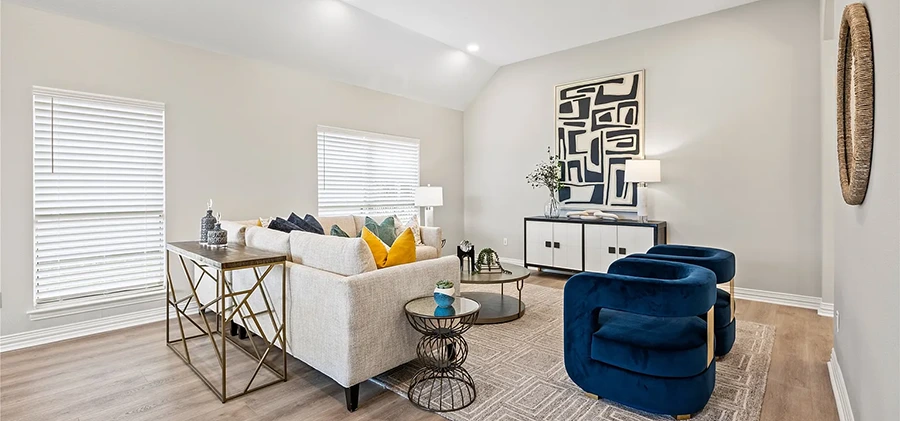 Wide view of a staged open living room featuring a geometric-patterned neutral rug, a beige sofa with yellow and blue pillows, two blue velvet accent chairs, and a geometric wood console table
