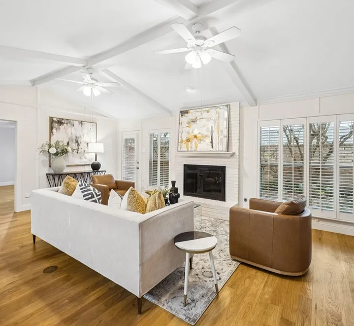 Bright living room staging with a cream sofa, cognac leather chairs, a white brick fireplace, and vaulted beam ceilings