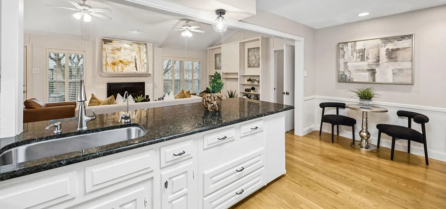Staged kitchen view showing a dark granite island sink looking into a living space and a small dining setup with black chairs