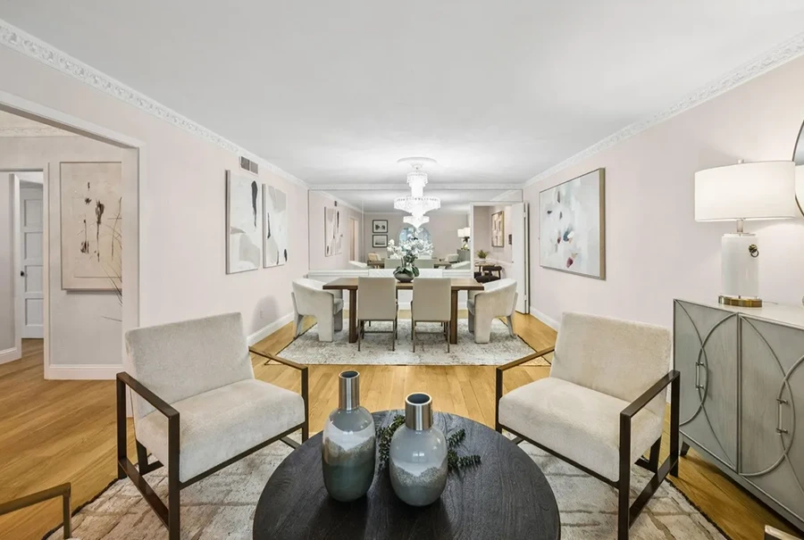A staged sitting area featuring two light grey upholstered armchairs with dark metal frames. In the background, a formal dining table with six cream chairs is set under a crystal chandelier